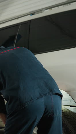 vista trasera de un ingeniero inspeccionando el neumático trasero de un jeep blanco usando la luz de una antorcha en una tienda de automóviles, con un colega en la parte delantera del vehículo, mostrando el mantenimiento del automóvil, el proceso de inspección de neumáticos