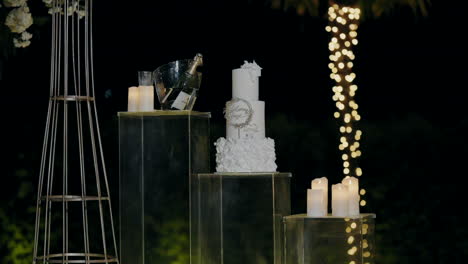 Elegante-Exhibición-De-Boda-Nocturna-Con-Un-Pastel-Blanco-Escalonado,-Champán-En-Una-Cubeta-De-Hielo-Y-Velas-Encendidas-Sobre-Pedestales-De-Vidrio