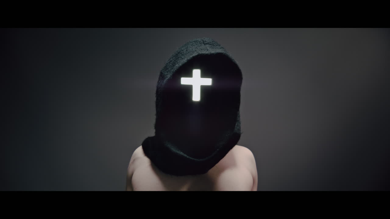 Masked and hooded man with cross of light glowing on his face rise his arms