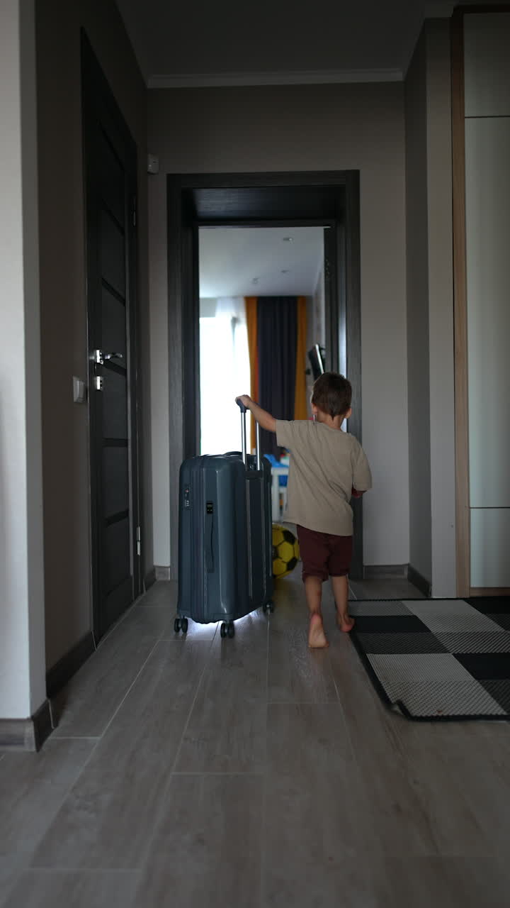 Little boy goes by the house pushing a suitcase. Vacation and travelling concept. Vertical video.
