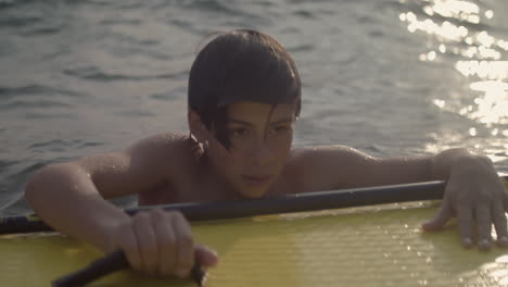 young man clutches paddleboard while floating in lake in thoughtful stare