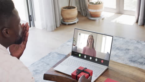 hombre afroamericano sosteniendo un regalo rojo y usando una computadora portátil con una mujer caucásica en la pantalla