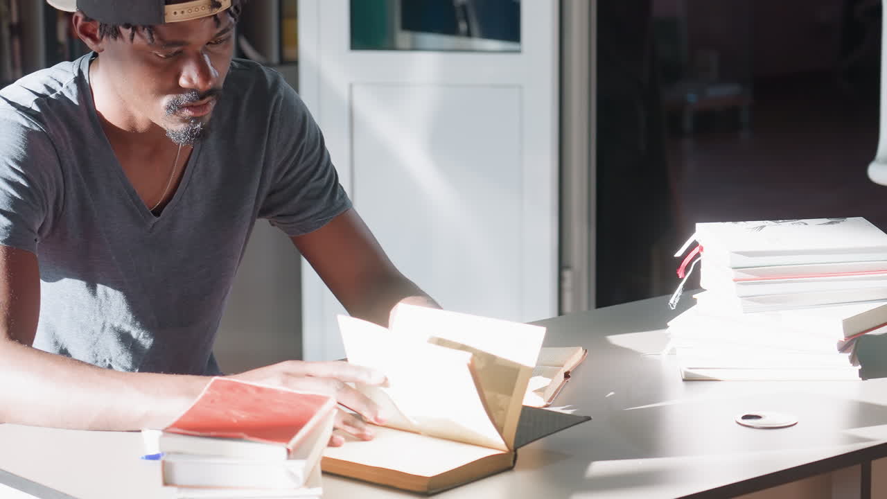 Student in cap prepares to study in bright library, seated by open book, arms stretched on table, eyes lifted toward window, calm focused mood, soft daylight and shelves behind