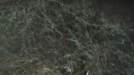vista aérea de arriba hacia abajo de un joven relajado con suéter rojo y jeans mira hacia el cielo tendido en un campo de hierba oscura, el aumento de drones revela bosque por carretera, día