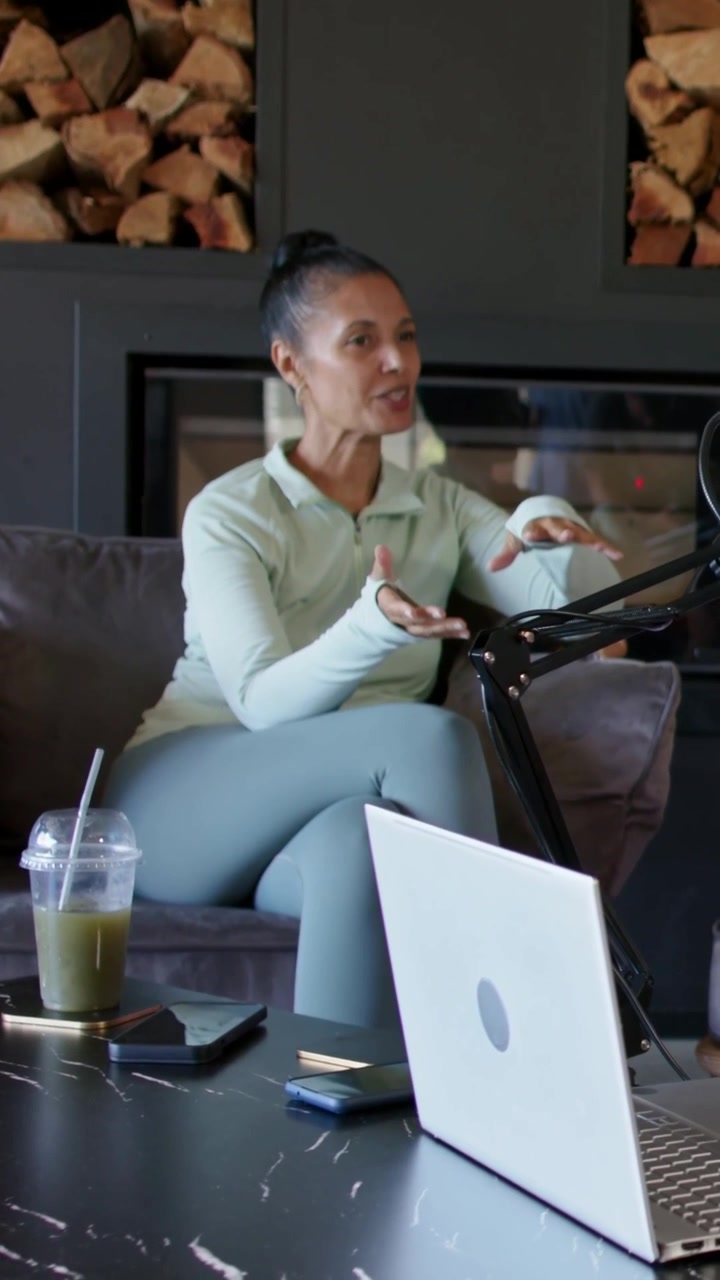 Vertical video: Speaking woman in mint set gesturing as cam rolling for podcast on couch with mic