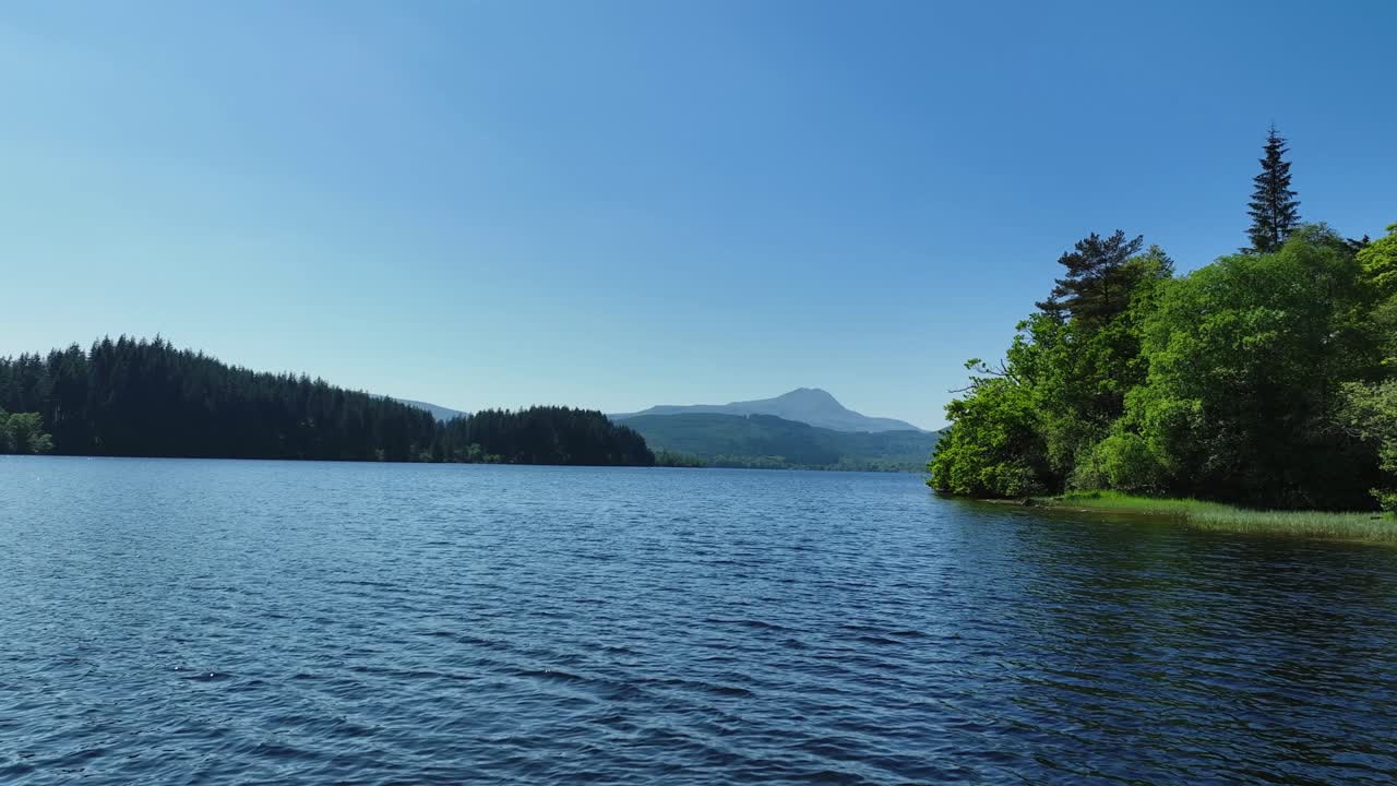 Aerial drone footage flying over the surface of still water of Loch Ard in the Troasachs National Park Scotland towards a native scots pine forest with clear blue sky on a sunny day