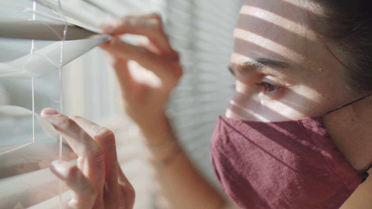 Woman in Mask Opening Looking through Window with Blinds