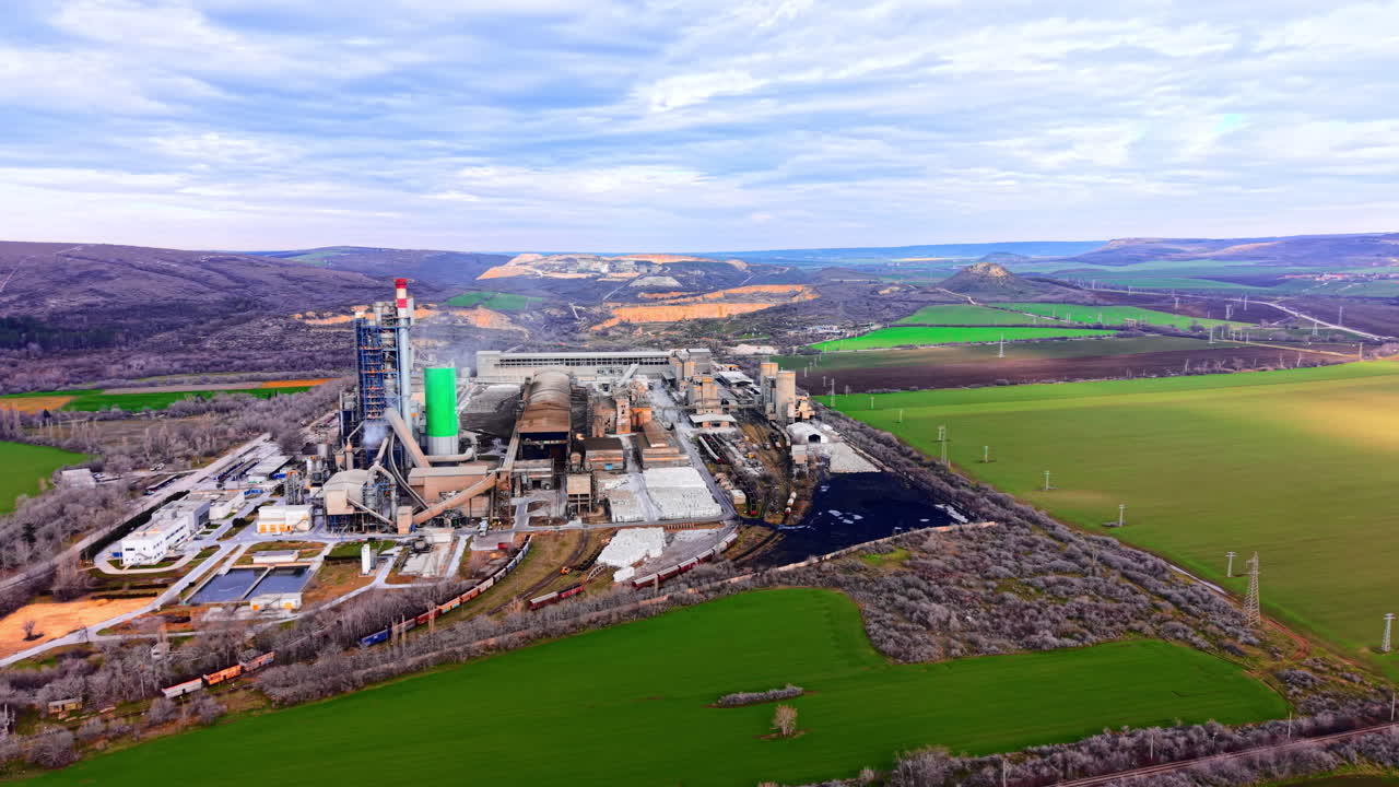 Planta de fabricación de cemento. Se observan silos de almacenamiento, naves de procesamiento y una vía férrea de carga. Instalación industrial con líneas de producción y vías férreas.