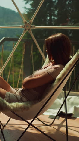 depressed woman sits in armchair with fur near panoramic window in tourist area overlooking dense forest and lake. relaxation in solitude and peace with nature