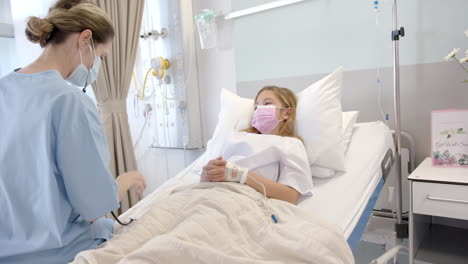 caucasian female doctor using stethoscope and girl patient in bed in face masks, slow motion