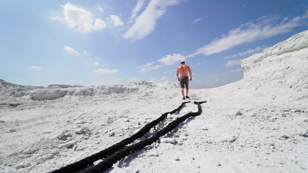 Shirtless man in shorts going up after his workout with ropes outdoors. Back view of a strong athlete rising on white mountain.