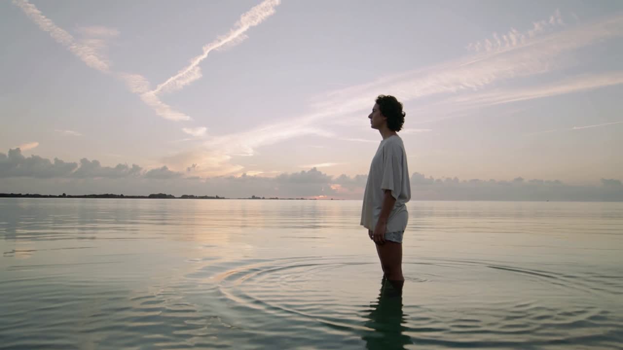 Uomo con camicia chiara e pantaloncini che osserva l'alba con l'acqua all'altezza della vita sulla riva, mentre le increspature si espandono.