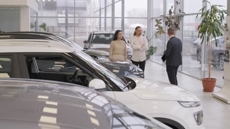 young car salesman showing to young couple new automobile at dealership salon.