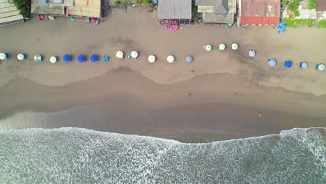 top down aerial view of batu belig beach coastline, row of parasols in soft sand, cafe buildings along winding road in bali, indonesia