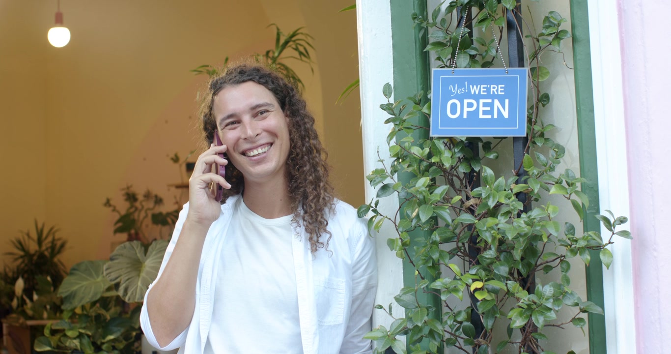 Persona adulta africana non binaria che risponde a una chiamata del negozio, controllando un telefono con custodia rosa, in piedi vicino al cartello 'aperto'.