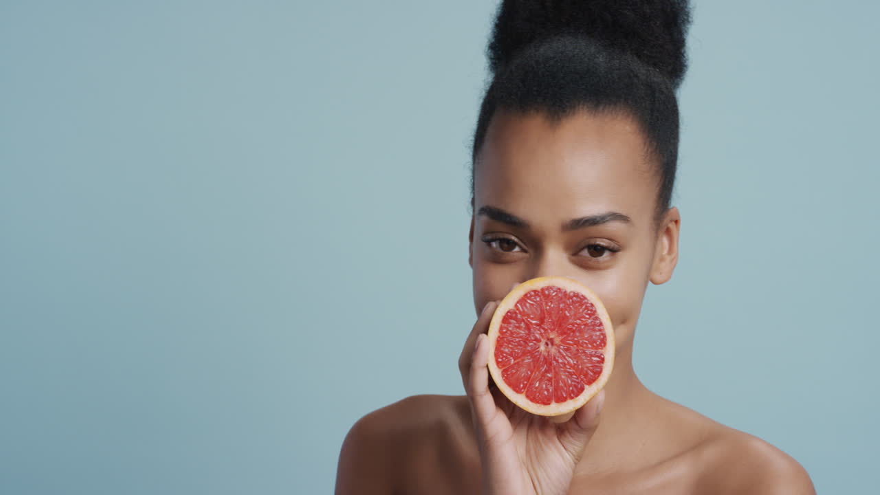 ritratto attraente giovane donna afroamericana che tiene in mano un pompelmo sorridendo godendo di un'essenza naturale di cura della pelle sana donna bella con una carnagione perfetta sullo sfondo blu