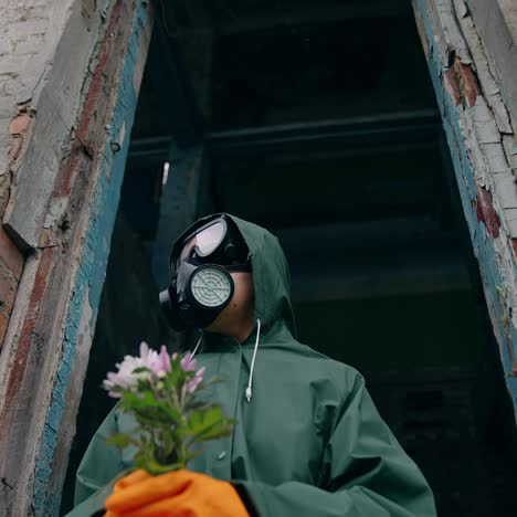 man with gas mask holding flowers. person in safety suit with flowers sitting in the abandoned building. ecology pollution concept. consequences of chemical attack
