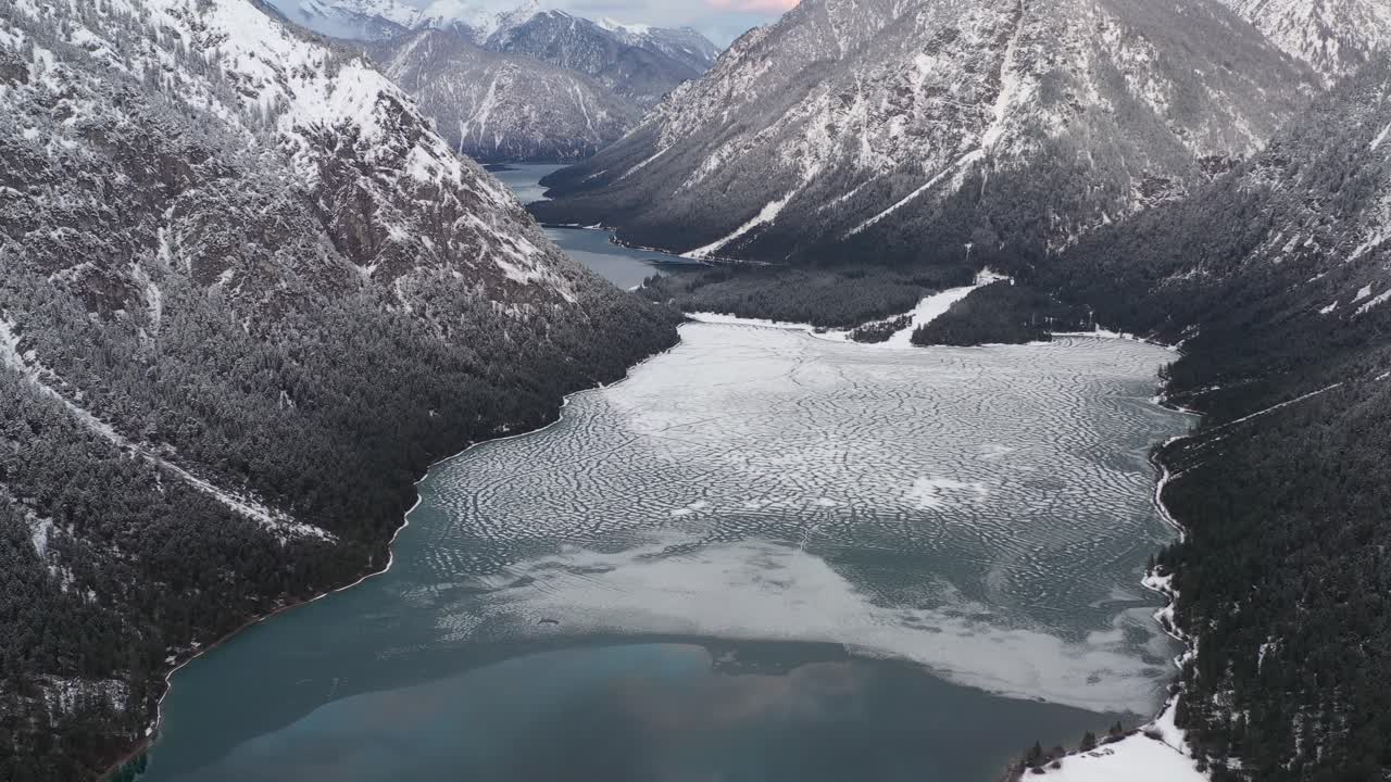 Vista aerea di un lago parzialmente ghiacciato tra catene montuose innevate.