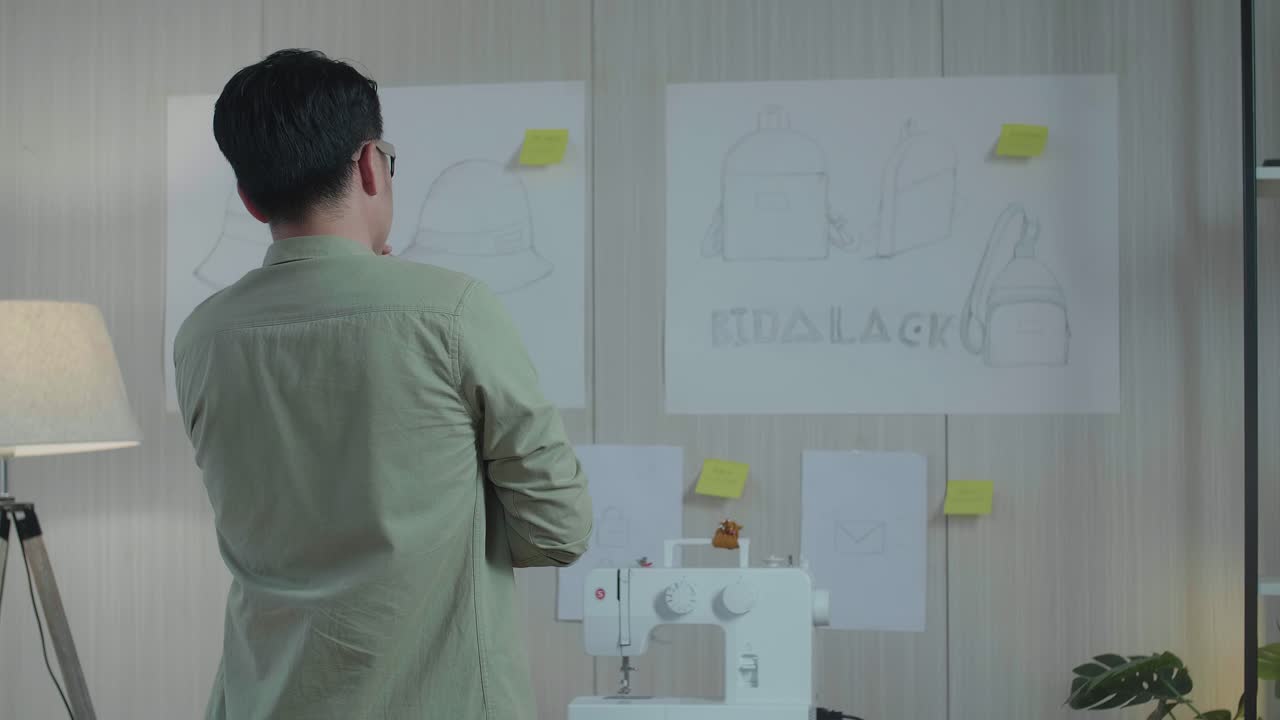 벽에 있는 가방 디자인 스케치 종이를 바라보는 아시아 남자 디자이너의 뒷면