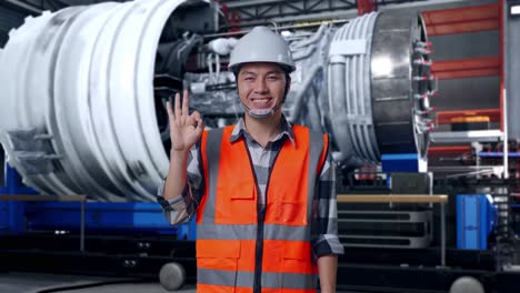 asian male engineer with safety helmet smiling and showing okay gesture to the camera while standing with airplane engine maintenance conducted, analytics checking the turbine