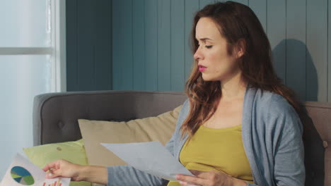 macro serious pregnant woman working with papers on couch in living room.