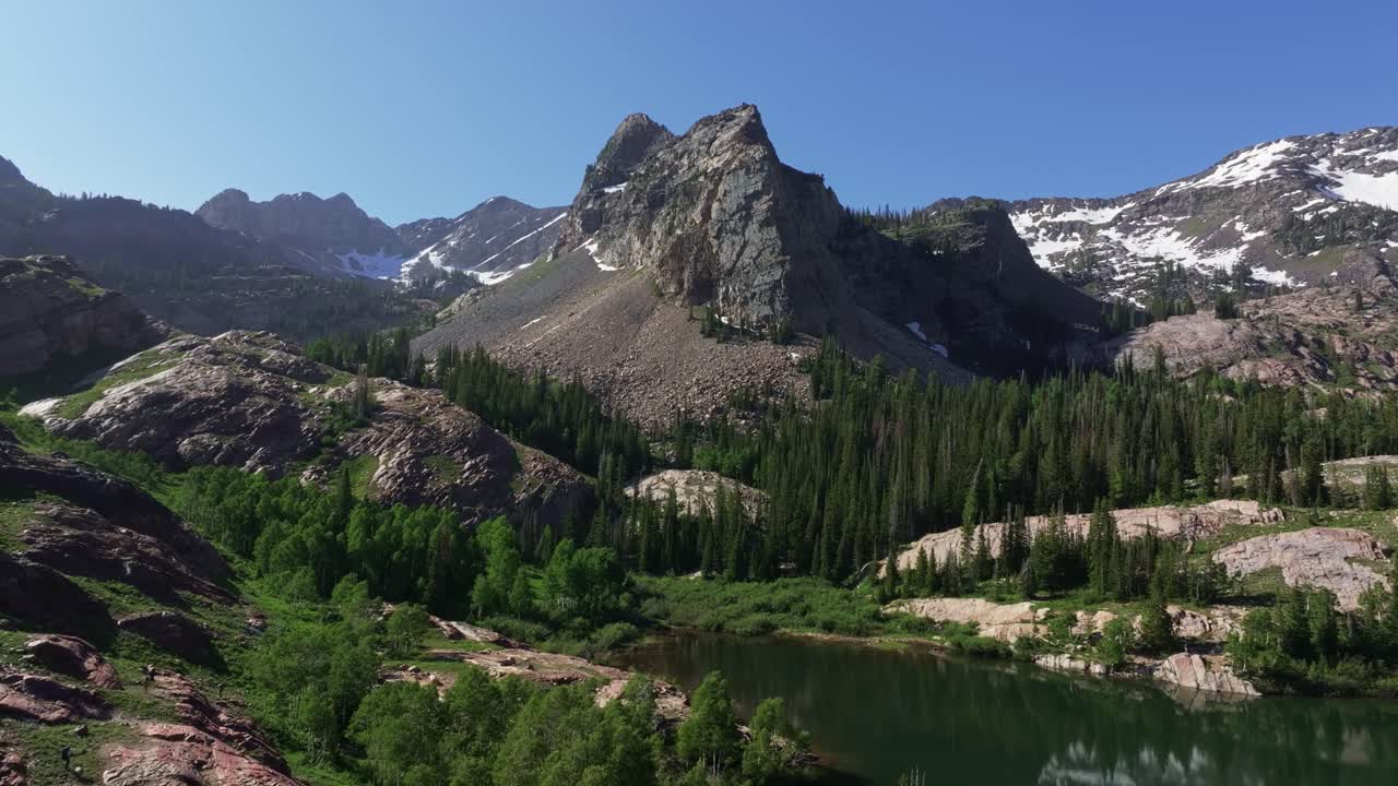 Aerial drone dolly out wide shot of Lake Blanche in Big Cottonwood Canyon near Salt Lake City, Utah, on a bright green summer morning with rocky peaks, lush foliage and Sundial Peak towering above