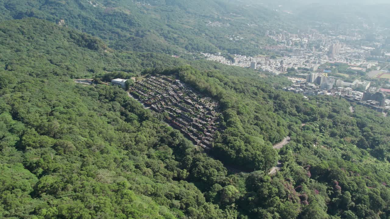 un cementerio tradicional taiwanés ubicado en un bosque verde exuberante con el paisaje urbano en el fondo, aéreo