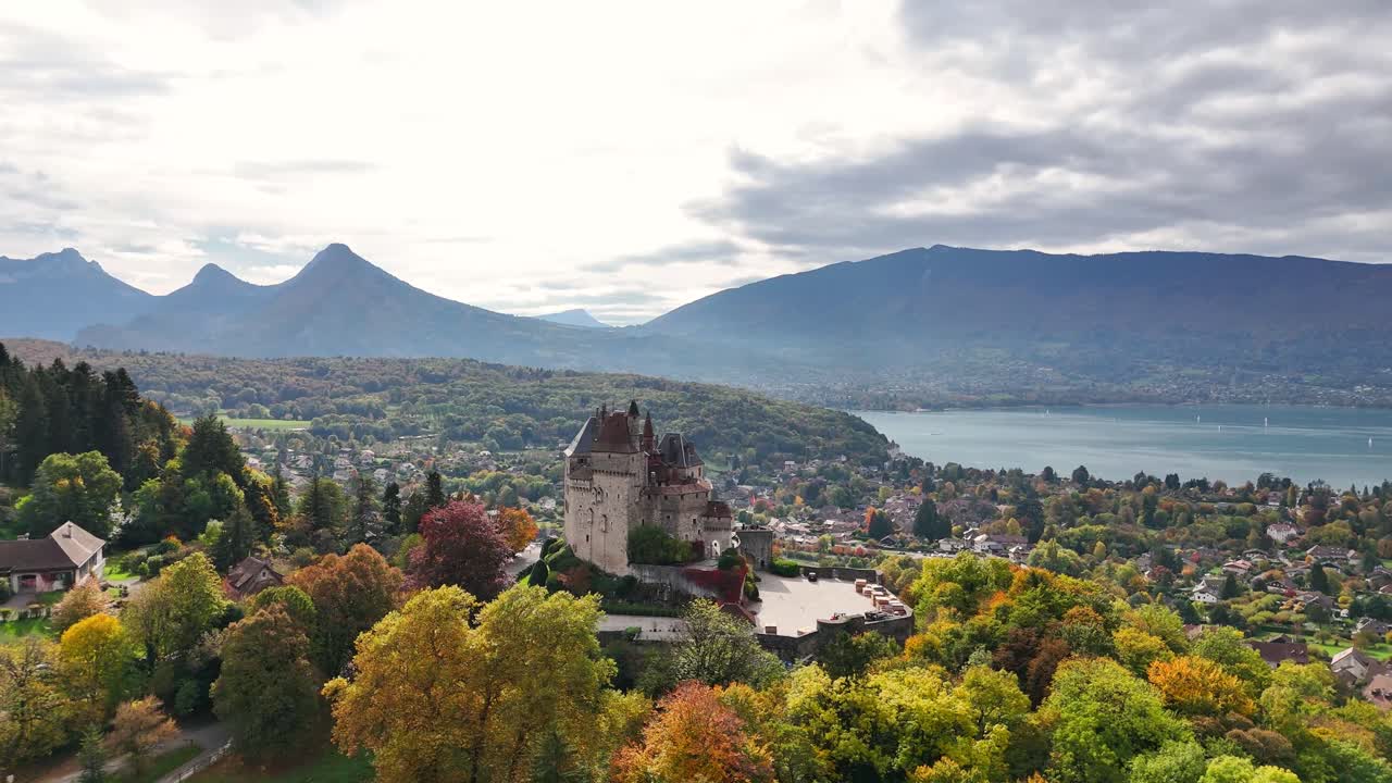 Château de Menthon‑Saint‑Bernard towers, courtyards and terraced gardens framed by autumn woodland, the village of Menthon‑Saint‑Bernard, blue waters of Lake Annecy and distant Alpine peaks aerial