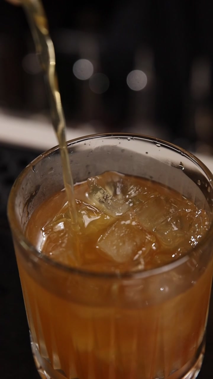 Primo piano del versamento di un cocktail ambrato in un bicchiere con ghiaccio