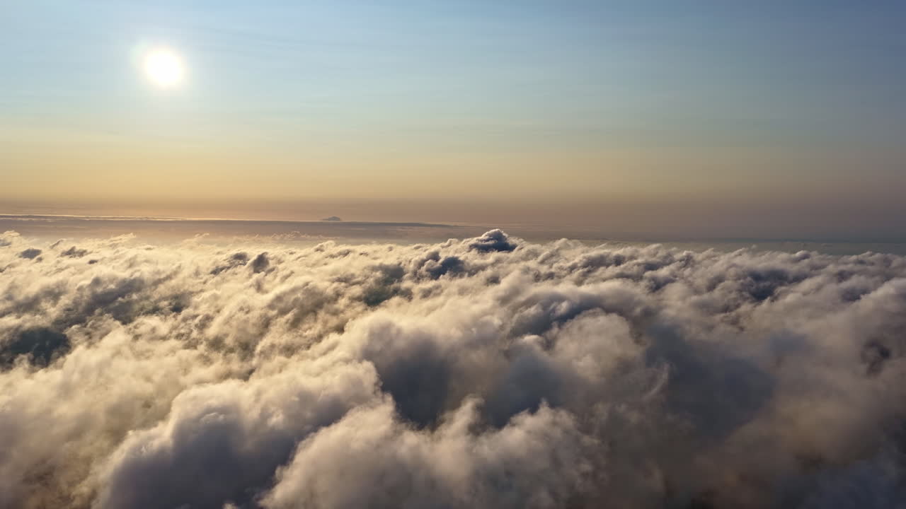Una serena scena aerea che mostra il sole che sorge sopra una fitta coltre di nuvole ondeggianti. Una luce dorata tenue si diffonde nel cielo mentre le cime delle nuvole si muovono delicatamente creando un'atmosfera sognante e pacifica.