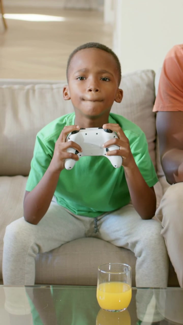 Vertical video: Game starting, child pressing buttons on pad on couch, wearing green tee, juice