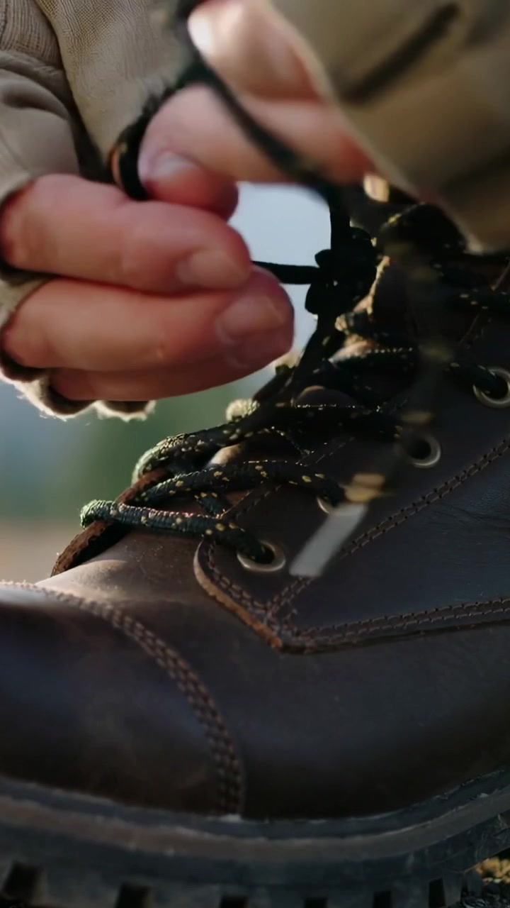 Vertical video: Tying beige-gloved hands securing laces on hiking boot on trail, preparing to hike