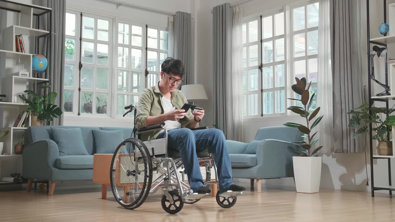 Asian Man Sitting In A Wheelchair While Use Mobile Phone And Credit Card Shopping Online At Home