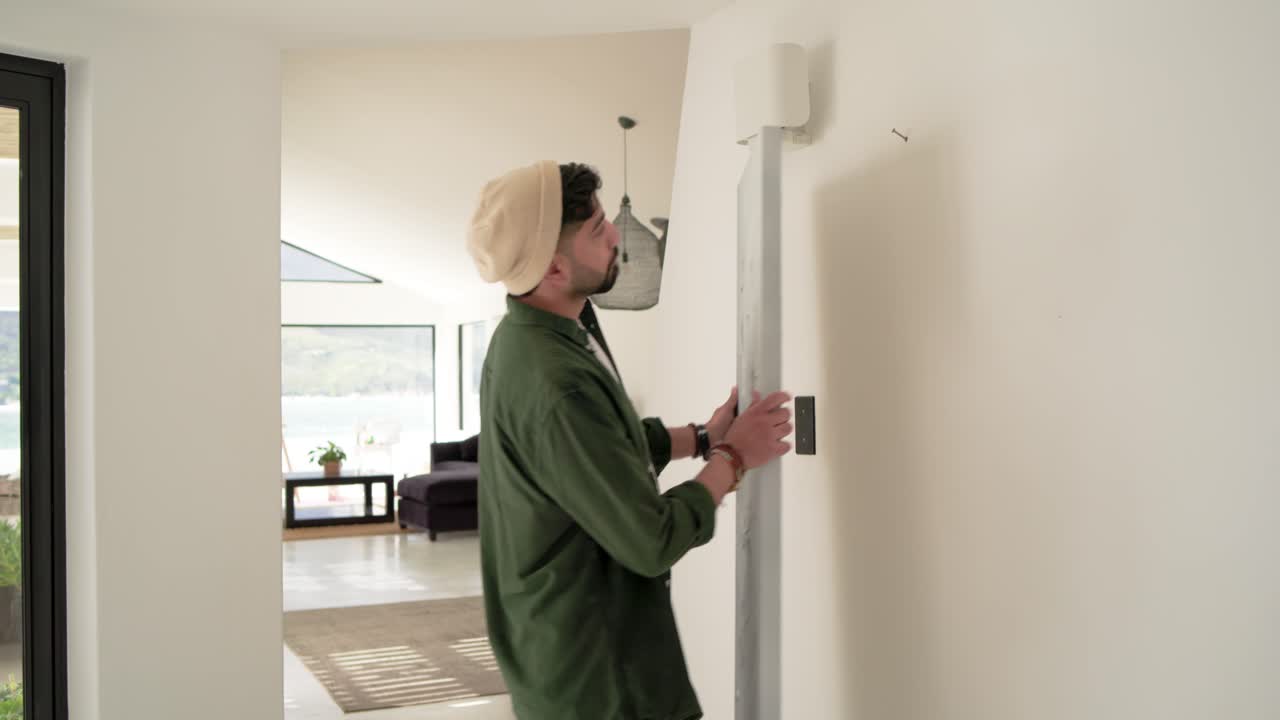 Testing canvas, Indian man in beanie lifting aligning canvas to hang on wall hook in hallway