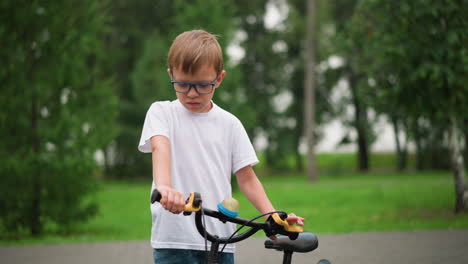 a young boy with glasses, focused and intent, blows something off his hand while keeping his other hand on the bicycle handlebar, he is wearing a white shirt