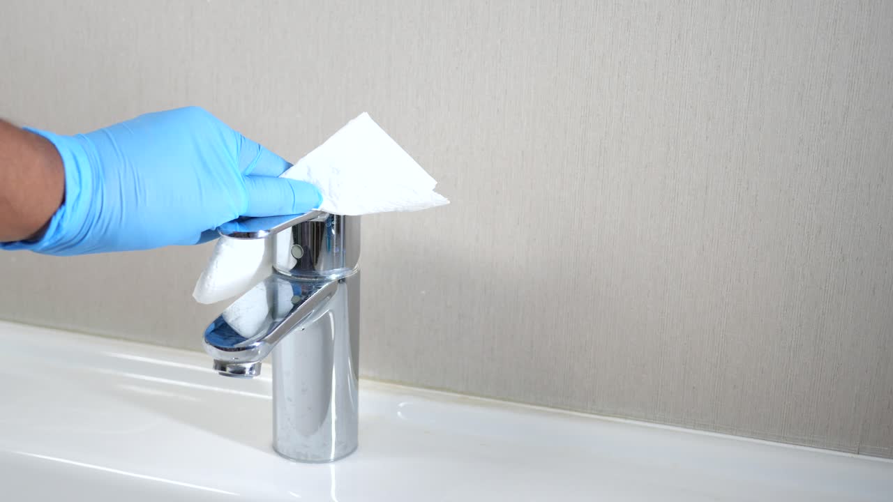 la mano de una persona con guantes azules limpia un grifo de cromo con una toalla de papel