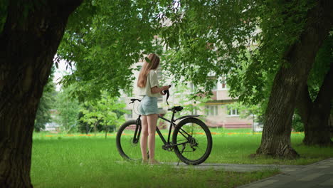 lady approaches black bicycle parked on paved path, puts on gloves on seat and hand, background shows a child running in distance, surrounded by tall trees, and residential buildings