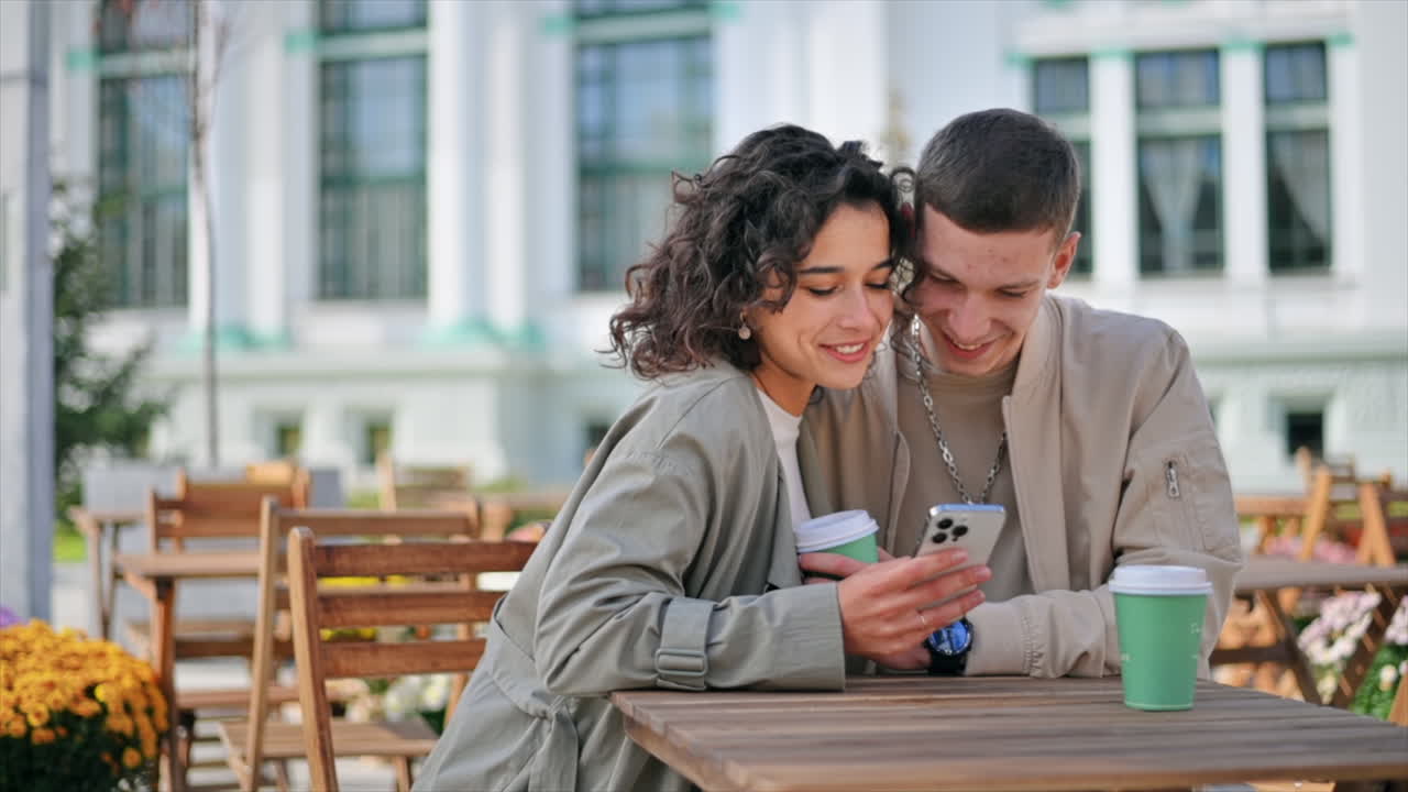 Una coppia felice all'aperto vicino a un bar. Che guarda il telefono, sorride, parla, beve caffè. Atmosfera autunnale. Rallentatore