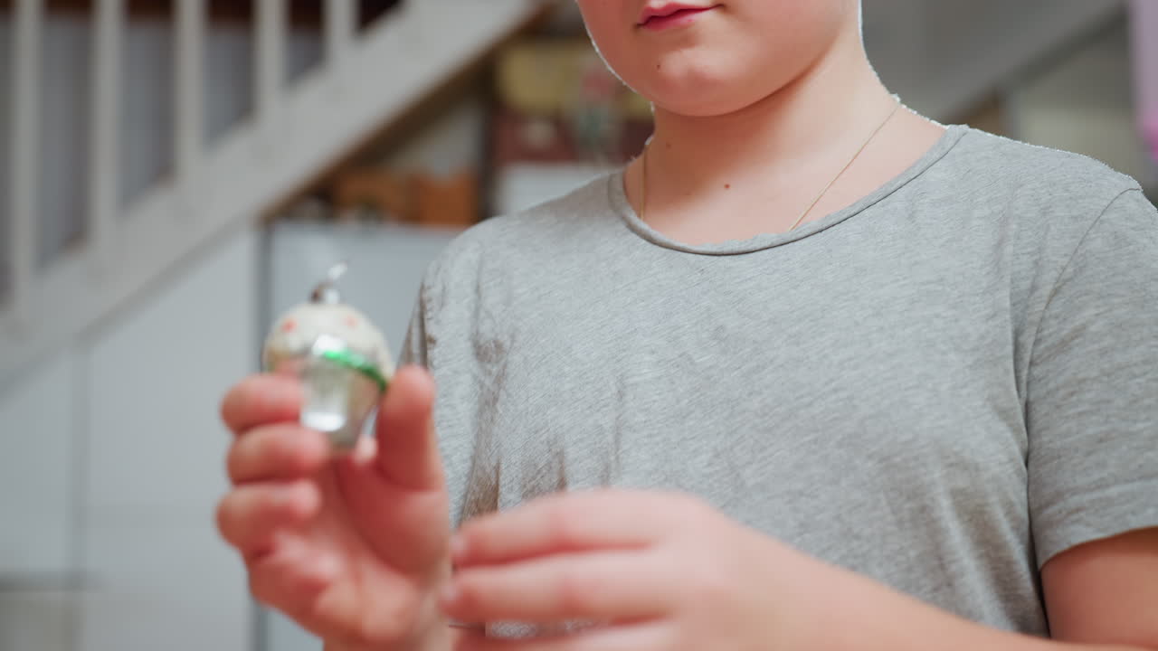 Close view of boy in grey top focusing while playing with blurred ornament in cozy home interior decorated with soft lighting and festive atmosphere, expressing concentration and quiet joy