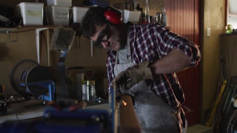 hombre caucásico fabricante de cuchillos en un taller con gafas y auriculares usando una lijadora