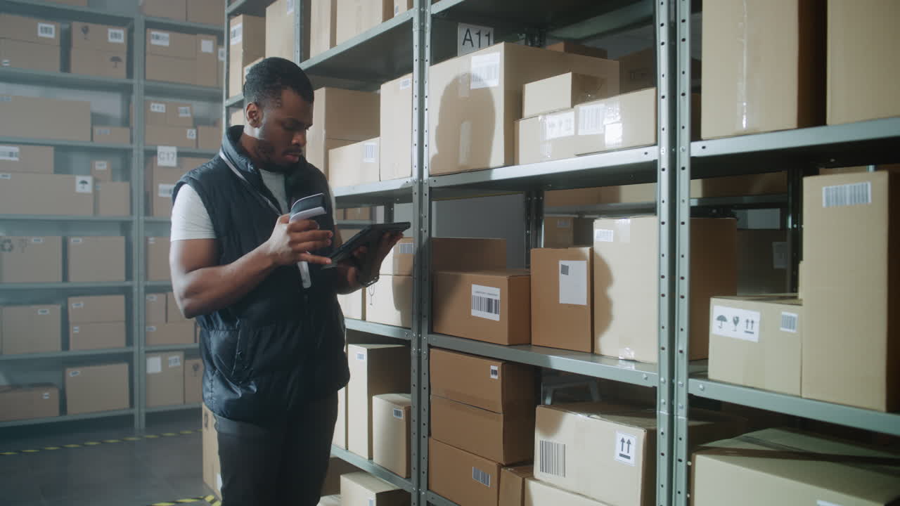 African American Inventory Specialist Walks in Warehouse Retail Facility Scans Parcels African American Inventory Specialist Walks in Warehouse Retail Facility Scans Parcels with Ecommerce Online Orders Checks Delivery Information on Tablet Computer Sorting Center of Postal Service