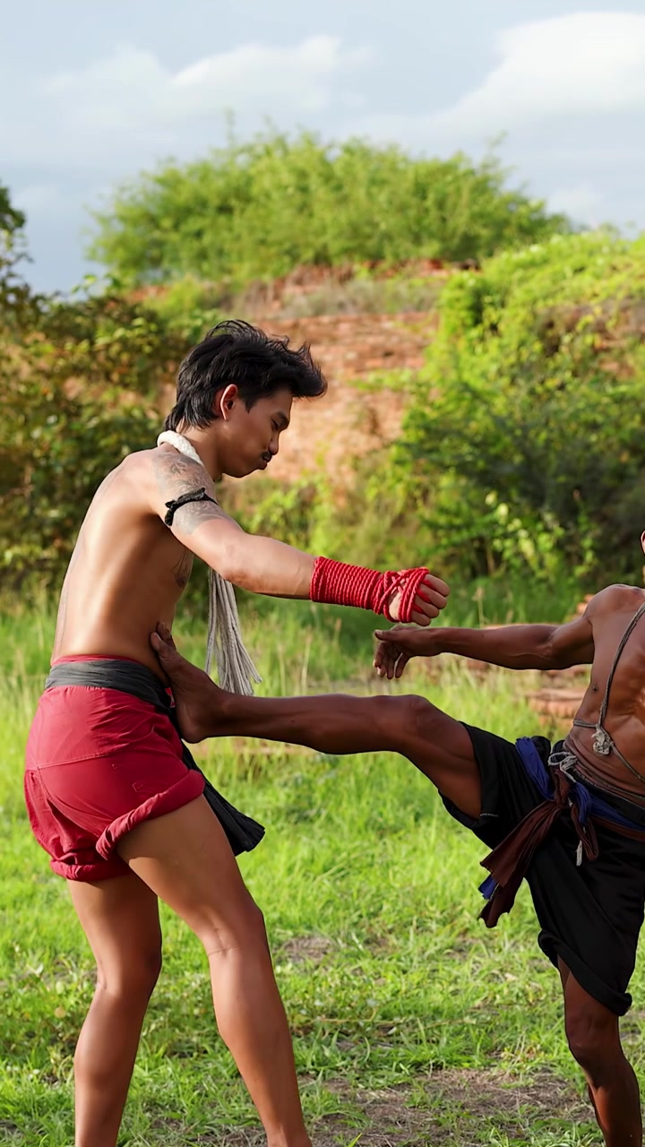 Athletic men practice Muay Boran combat techniques outdoors near historic brick ruins in daylight.