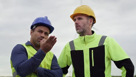 caucasian and latin male engineers standing on wind turbine field and discussing together.