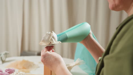 baker uses large green piping bag to apply white icing onto dessert base held with metal tool during decorating process while sitting at table with swirl cupcakes and parchment paper in cozy kitchen