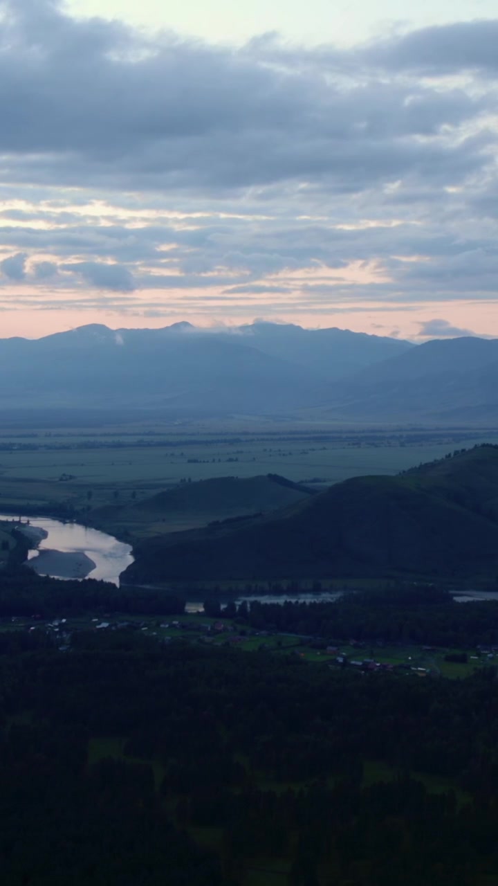 Riprese aeree con drone di un ampio paesaggio dei Monti Altai con un fiume tortuoso, colline boscose e vette distanti sotto nuvole imponenti al crepuscolo, Siberia, Russia.