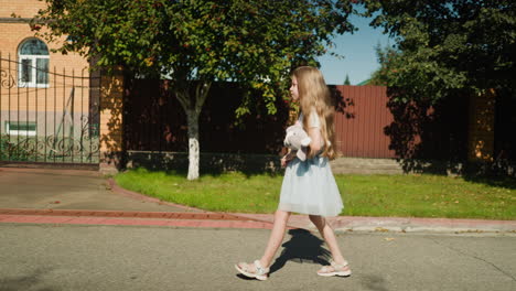 side view of young girl in blue gown with net holding stuffed teddy bear walking along quiet neighborhood street as sunlight casts clear shadow on ground with trees and fence in background