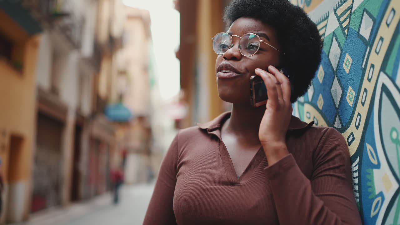 chica africana atractiva hablando por teléfono inteligente, pareciendo feliz al aire libre