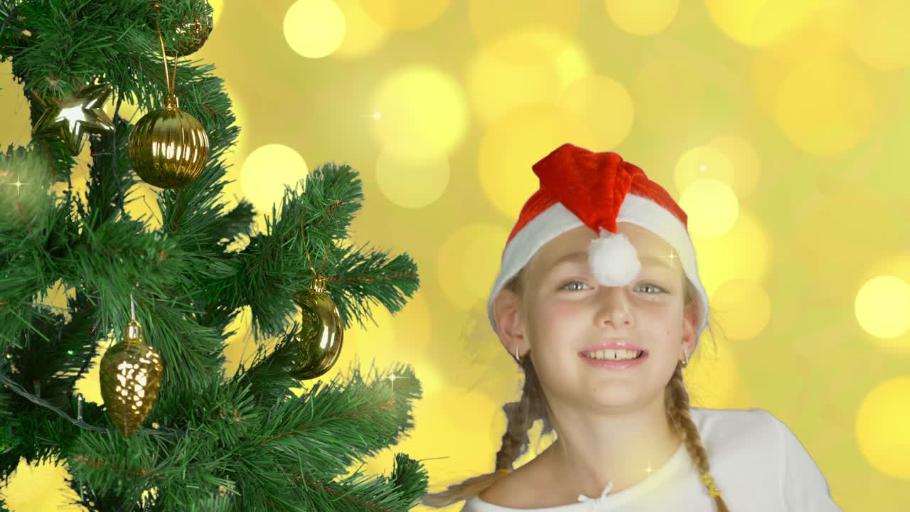 Happy young girl dancing near christmas tree in Santa red hat. Happy young caucasian girl indoors studio close up shot. Golden background. Decorated christmas tree.