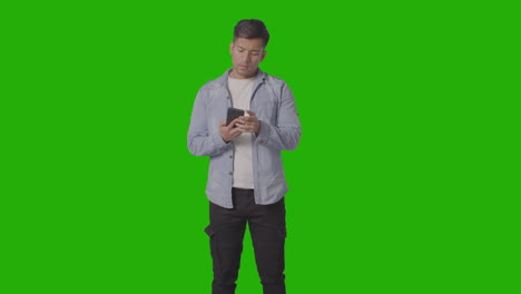 Studio-Shot-Of-Casually-Dressed-Young-Man-Checking-Mobile-Phone-Against-Green-Screen-