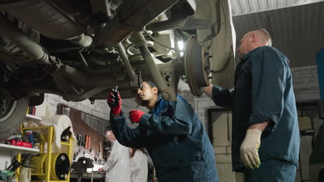 mecánico trabaja bajo el coche usando una llave mientras un colega sostiene la luz para una mejor visibilidad, dos técnicos de laboratorio se ven trabajando en el fondo en un taller mecánico